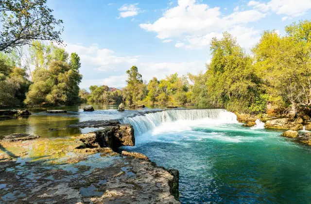 De brede Manavgat waterval met turquoise water en groene oevers onder een blauwe lucht